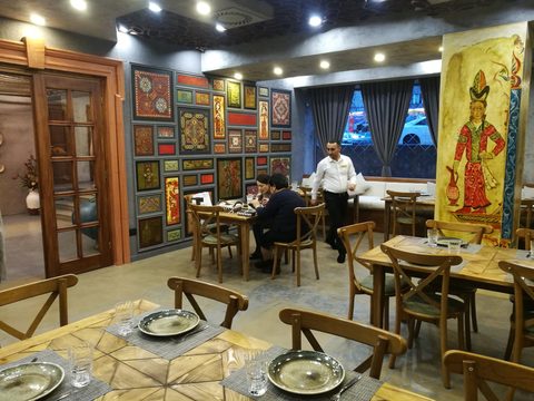 Restaurant interior: a wall covered with framed painted panels of carpets, manuscripts and reliefs, and a column painted with a woman holding a jug