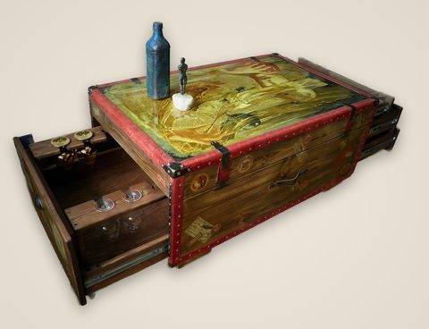 Trunk Table: a wooden coffee table made like an old travelling trunk, edged in red with brass studs and covered with old luggage labels, its lid painted with a woman holding a red sphere, a bar drawer with glasses pulled out at one end