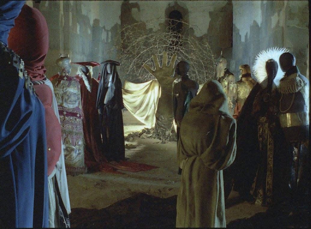 Film set: Robed and costumed figures gather around a giant stone hand in a ruined hall.